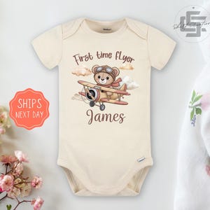 May include: Cream-colored baby onesie with a cartoon illustration of a teddy bear in a pilot's hat and scarf, flying a biplane. The text "First time flyer" and the name "James" are printed on the onesie.