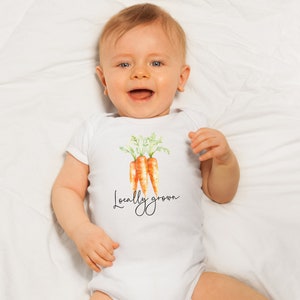 May include: A white baby onesie featuring a watercolor illustration of three carrots with green tops. The text "Locally grown" is written in a cursive font below the carrots. The baby is lying on a white sheet.