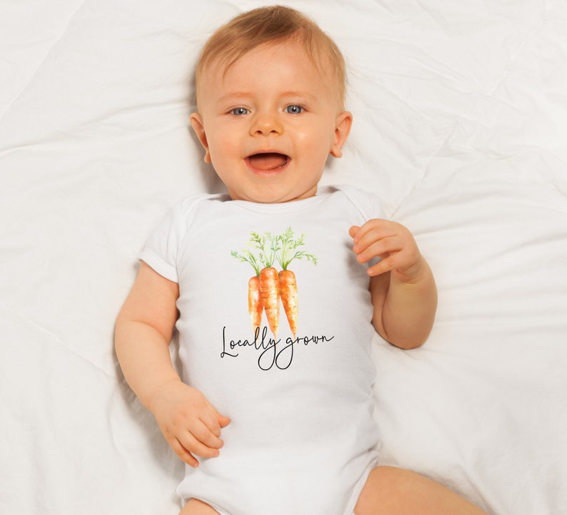 May include: A white baby onesie featuring a watercolor illustration of three carrots with green tops. The text "Locally grown" is written in a cursive font below the carrots. The baby is lying on a white sheet.