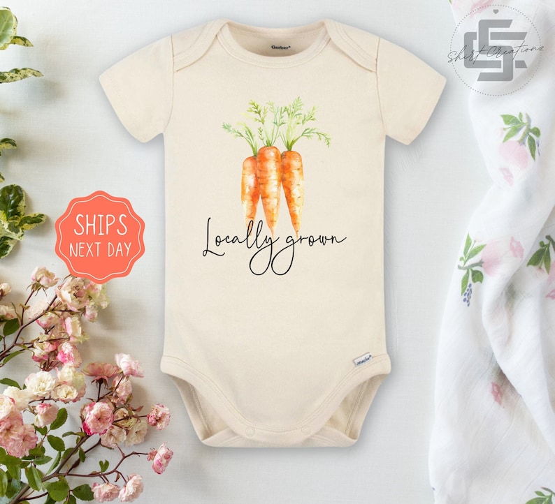 May include: Cream-colored baby onesie featuring a watercolor illustration of three carrots with green tops. The words "Locally grown" are written in a cursive font below the carrots. The onesie is made of soft material.