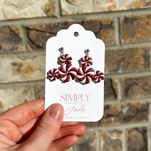 Może przedstawiać: Kolczyki w kształcie czerwono-białych cukierków miętowych. Prezentowane na białej karcie z napisem "SIMPLY Jade" i "HANDMADE CLAY EARRINGS". Kolczyki wykonane są z gliny i mają błyszczące wykończenie.