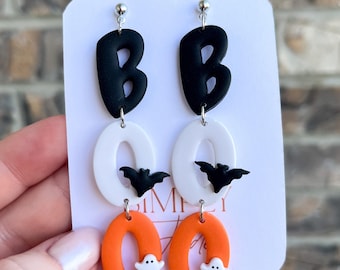 May include: Halloween-themed earrings with a playful design. The earrings feature black letter "B" shapes, white oval shapes with black bat accents, and orange oval shapes with white ghost charms. The earrings are attached to a white card.