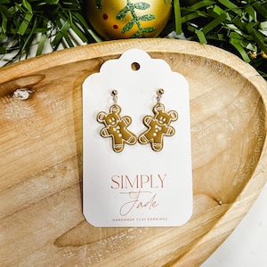 Może przedstawiać: Złote kolczyki w kształcie ludzików z piernika z białymi detalami lukru, prezentowane na białej karcie. Na karcie napis "SIMPLY Jade HANDMADE CLAY EARRINGS". Tło przedstawia drewnianą tacę, zieloną girlandę i złotą kulę.