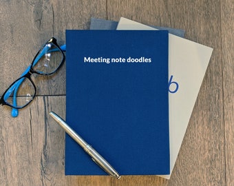 May include: A blue notebook with the text "Meeting note doodles" on the cover. The notebook is stacked on top of two other notebooks, one gray and one beige. A silver pen and blue-framed glasses are on a wooden surface.