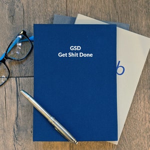 May include: A blue notebook with the text "GSD Get Shit Done" on the cover. The notebook is stacked on top of two other notebooks. A pen and glasses are on the wooden surface.