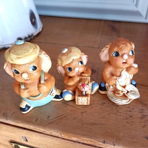 Könnte beinhalten: Drei Vintage-Hasenfiguren aus Keramik. Die Hasen sind braun mit blauen Augen und tragen Hüte und Kleidung. Ein Hase hält ein Tuch mit blauen Punkten. Die Figuren stehen auf einer Holzoberfläche.