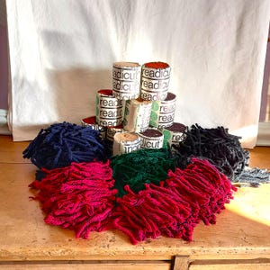 Könnte beinhalten: Eine Sammlung von Garnen in verschiedenen Farben, darunter Rot, Grün, Blau und Schwarz, auf einer Holzoberfläche angeordnet. Mehrere kleine, zylindrische Behälter mit dem Aufdruck "readicut" sind im Hintergrund gestapelt.
