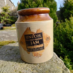 Puede incluir: Un tarro de mermelada antiguo de cerámica con borde marrón y etiqueta descolorida. La etiqueta dice "UNECOL FULL FRUIT STANDARD JAM". El tarro tiene forma cilíndrica y es de color beige claro. El tarro está sobre una superficie de piedra.