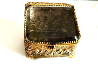 May include: A square gold-toned metal box featuring intricate relief carvings of figures and animals on the sides. The lid is made of a thick, beveled piece of dark, translucent glass with a serrated metal border framing the top edge.