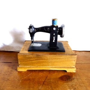 May include: A miniature black sewing machine model with gold-toned decorative scrollwork, a silver-coloured handwheel, and a light blue thread spool mounted on top. The machine is securely attached to a rectangular, light brown wooden base.