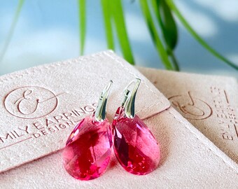 May include: A pair of silver dangle earrings with pink crystal teardrop shaped stones. The earrings are displayed on a pink velvet pouch with the text "Emily Earrings - Design" printed on the front.