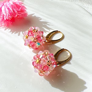 Può includere: Un paio di orecchini rosa e dorati con un gruppo di perline rosa a forma di fiore. Gli orecchini sono su una superficie bianca.