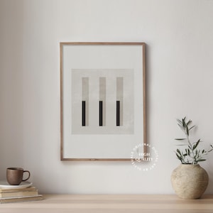 Puede incluir: Una impresión abstracta enmarcada con tres barras verticales en tonos beige y negro. La impresión está en un marco de madera claro y está colgada en una pared blanca.