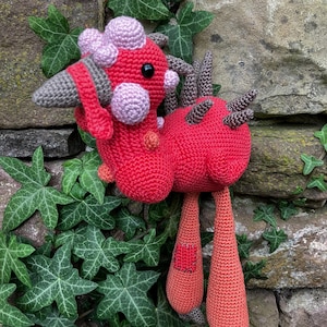 Puede incluir: Una criatura de ganchillo roja hecha a mano con detalles en rosa, gris y naranja. El diseño caprichoso presenta un cuerpo redondeado, cuernos y largas patas colgantes. La criatura se encuentra sobre un fondo de hiedra verde y una pared de piedra.