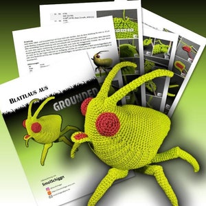 May include: A crocheted green insect with red circular eyes and long antennae is displayed next to printed instructions. The text "BLATTLAUS AUS GROUNDED" is visible on the paper. The insect is a handmade craft item.