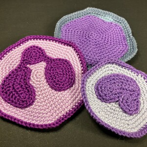 Può includere: Sottobicchieri all'uncinetto a forma di cellule, con disegni viola e lavanda. I sottobicchieri hanno un aspetto dettagliato e strutturato, con un focus sulle strutture interne delle cellule. Il set include tre sottobicchieri, ognuno con una rappresentazione cellulare unica.