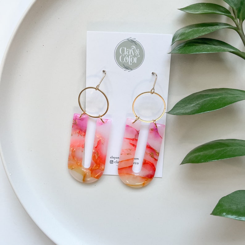 May include: A pair of pink and orange resin earrings with gold hoops. The earrings are shaped like a U with a thin opening in the center. The earrings are handmade and have a unique, abstract design.