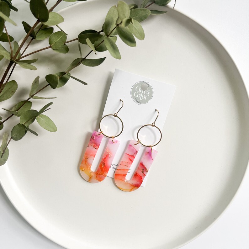 May include: A pair of pink and orange resin earrings with gold accents. The earrings are shaped like a U and have a gold hoop at the top. The earrings are on a white background with a card that says "Clay & Color".