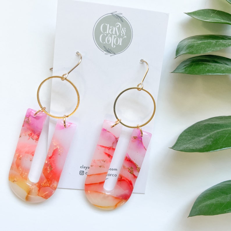 May include: A pair of gold hoop earrings with pink and orange resin U-shaped charms. The charms have a marbled effect with gold flecks.