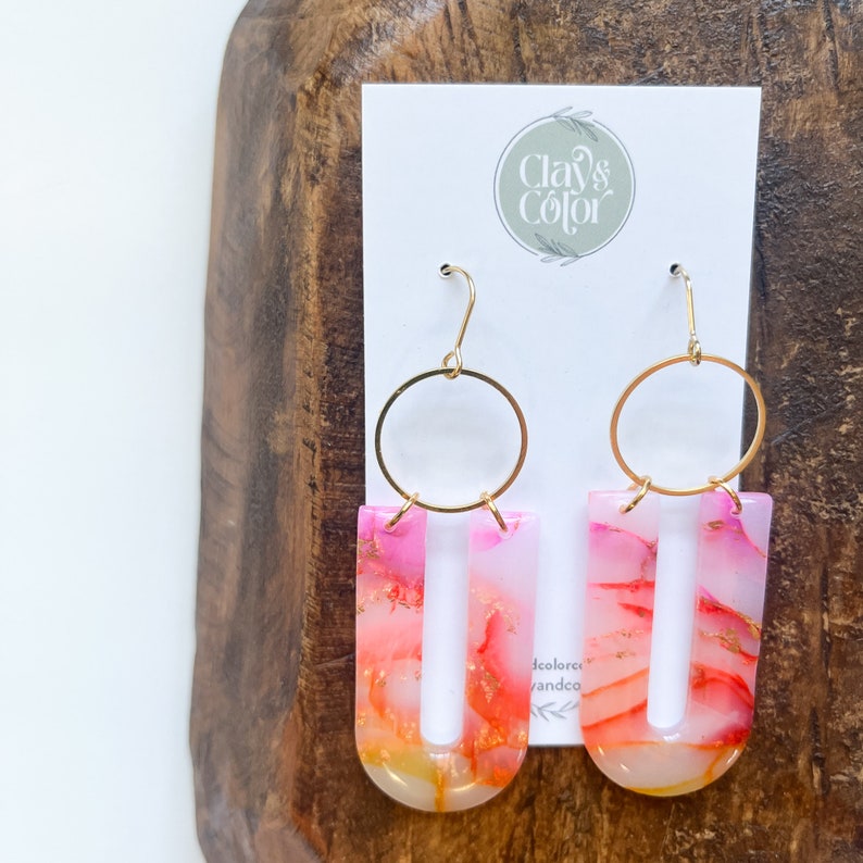 May include: A pair of pink and orange resin earrings with gold hoops. The earrings are shaped like a U with a thin opening in the center. The earrings are handmade and have a unique marbled effect.