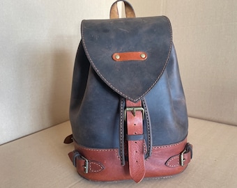 May include: A dark brown leather backpack with a brown leather strap and buckle. The backpack has a flap with a brown leather strap and buckle. The backpack has a brown leather bottom.