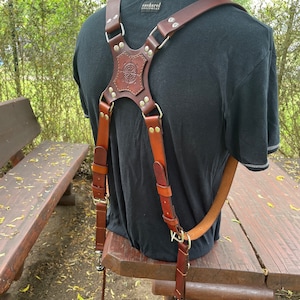 May include: Brown leather camera strap with a compass design and metal accents. The strap is worn over a person's shoulders and is secured with buckles.