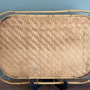 May include: A rectangular bamboo serving tray with a woven, light brown surface. The tray has a rounded frame and handles, and is supported by a black stand. The woven surface has a diagonal pattern.