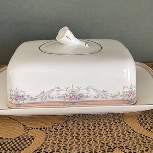May include: White porcelain butter dish with a lid and a matching tray. The dish features a floral and scroll design in pink, purple, and green, with gold trim. The lid has a curved handle. A decorative serving piece.
