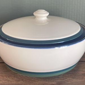 Puede incluir: Un cuenco de cerámica blanca con tapa. El cuenco tiene forma redondeada y está decorado con bandas azules y verdes alrededor del borde y la base. La tapa tiene un pomo blanco en la parte superior.