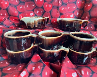 May include: A collection of brown ceramic soup bowls and a creamer with a drip glaze. The bowls have handles and are stacked in pairs. The creamer has a spout and a handle. The background is a red fabric with a cherry pattern.