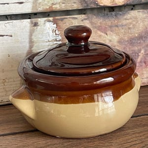 Puede incluir: Una pequeña cazuela de cerámica cubierta con un diseño de dos tonos. La tapa y la parte superior son de color marrón oscuro brillante, mientras que la sección inferior es de color beige claro. El plato tiene asas pequeñas.