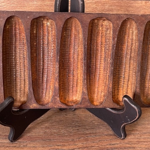 Może przedstawiać: Antyczna żeliwna forma do chleba kukurydzianego z sześcioma indywidualnymi formami. Forma jest zardzewiała i ma ciemnobrązowy kolor.