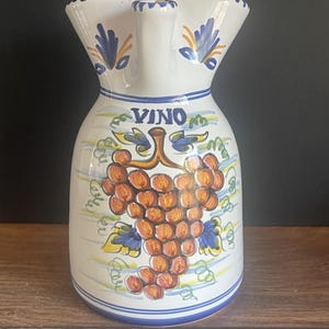 May include: White ceramic pitcher with a wide, scalloped rim and a handle. The pitcher features a hand-painted design of a bunch of grapes with the word "VINO" above it. Blue and yellow accents complete the design.