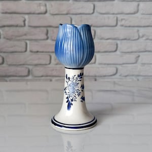 Może przedstawiać: Ceramiczny świecznik w kształcie tulipana z niebieskimi płatkami u góry i białą podstawą. Trzon ozdobiony jest niebieskim wzorem kwiatowym, a dolna krawędź posiada dwa niebieskie pierścienie. Przedmiot ma gładkie, błyszczące wykończenie.
