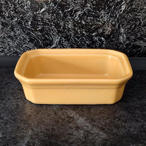 May include: A rectangular ceramic baking dish with a smooth, glossy finish in a solid light yellow colour. The dish features slightly angled sides and a thick, rounded rim, providing a simple and functional design for kitchen use.