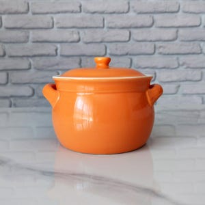Könnte beinhalten: Ein leuchtend orangefarbener Keramiktopf mit passendem Deckel und zwei seitlichen Griffen. Der Topf hat eine glatte, glänzende Oberfläche und eine abgerundete Form, die zum Kochen oder Servieren von Speisen dient. Der Deckel hat einen kleinen Knopf.