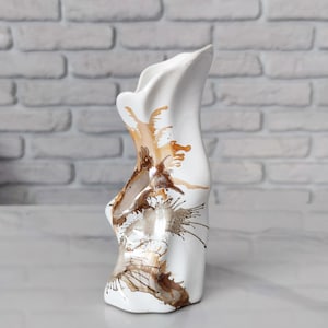 Könnte beinhalten: Eine weiße Keramikvase mit unregelmäßiger, wellenförmiger Silhouette und glänzender Oberfläche. Das Design zeigt abstrakte, fließende braune und hellbraune spritzartige Muster und Streifen, die sich von der Basis bis zum asymmetrischen Rand ziehen.