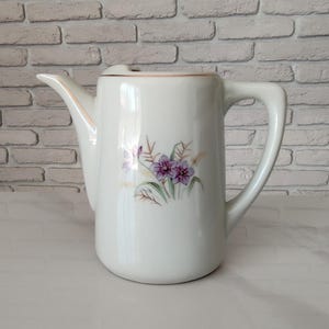 Op de afbeelding: Een witte keramische theepot met een verfijnde bloemenillustratie van paarse bloemen en groene stengels op de voorzijde. De theepot heeft een gebogen handvat, een puntige tuit en een dunne goudkleurige rand langs de bovenkant van de pot.