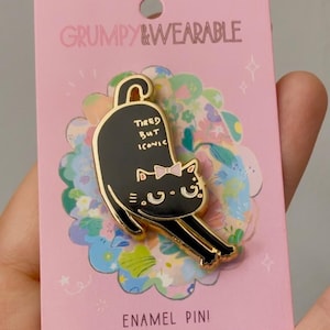 May include: A black enamel pin featuring a stretching cat with the text "Tired But Iconic" on a pink backing card. The card has the words "Grumpy & Wearable" and "Enamel Pin!" printed on it. The pin is set against a floral design.