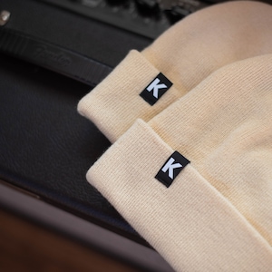 May include: Two beige knit beanies with a black 'K' logo on each.