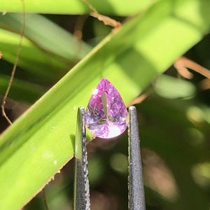 Op de afbeelding: Een peervormige, gefacetteerde edelsteen met een levendige roze en lichtpaarse tint. De doorschijnende steen wordt vastgehouden door een zilverkleurige metalen pincet, wat het heldere, reflecterende oppervlak en de precieze snit benadrukt.
