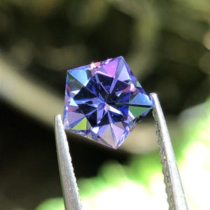 Op de afbeelding: Een vijfhoekige edelsteen met een diepe violette en blauwe kleur, voorzien van complexe geometrische facetten die het licht reflecteren. De steen wordt vastgehouden door een metalen pincet, wat de scherpe, symmetrische randen en kristalstructuur toont.