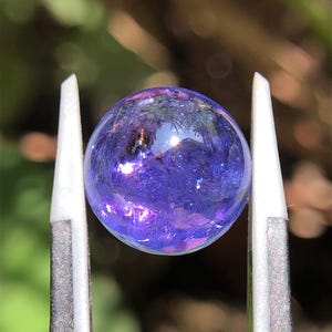 May include: A round, translucent tanzanite gemstone with a deep violet-blue hue, held securely between the two metal tips of a pair of tweezers. The stone exhibits internal light reflections and a smooth, polished surface texture throughout.