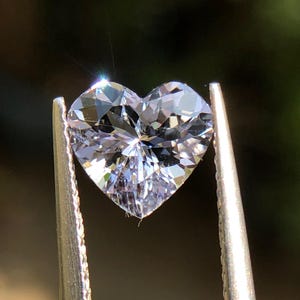 May include: A heart-shaped, faceted gemstone held by silver tweezers. The clear gem sparkles with multiple facets, reflecting light. The background is a blurred, dark green.