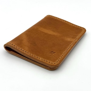 May include: A rectangular, tan leather wallet with a stitched border. The wallet has a minimalist design with a small, embossed logo on the front. The leather has a natural, slightly distressed appearance.