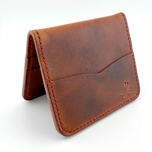 May include: A brown leather wallet with red stitching. The wallet has a curved pocket on the front and a logo in the lower right corner. The wallet is open, revealing card slots.