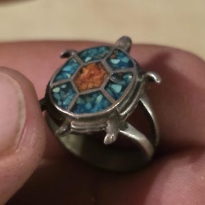 May include: A silver-toned ring featuring a turtle-shaped centerpiece. The shell is inlaid with small, fragmented pieces of bright blue turquoise and a central cluster of orange coral, set within a segmented metal frame with four legs.