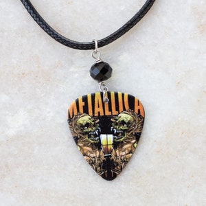 Puede incluir: Collar de cordón de cuero negro con una cuenta negra y un colgante de púa de guitarra con un logotipo de Metallica y un gráfico de calavera.