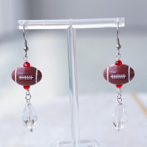 May include: A pair of dangle earrings featuring a brown and white football charm with red accents and a clear crystal dangle.