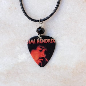 May include: A black necklace featuring a guitar pick pendant with a portrait of Jimi Hendrix. The pendant is black with a red and orange image of the musician. A black bead and a silver-colored metal loop connect the pendant to the black cord.